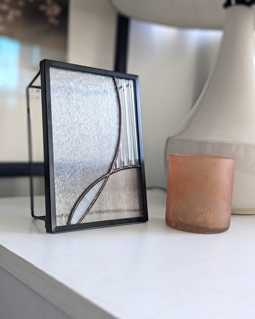 Decorative glass panel with black frame on a white surface, photographed from the left