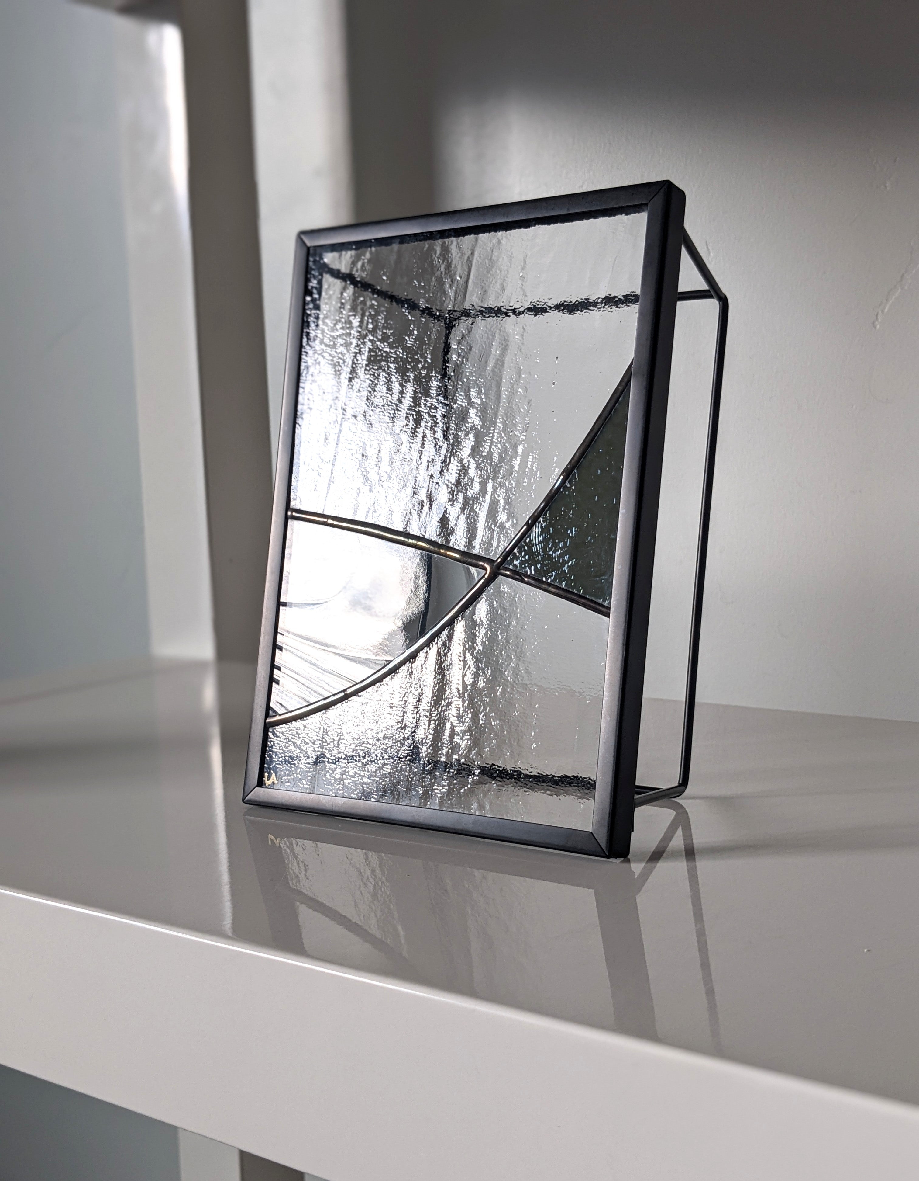 Modern black-framed glass artwork on a white shelf with shadows in the background.