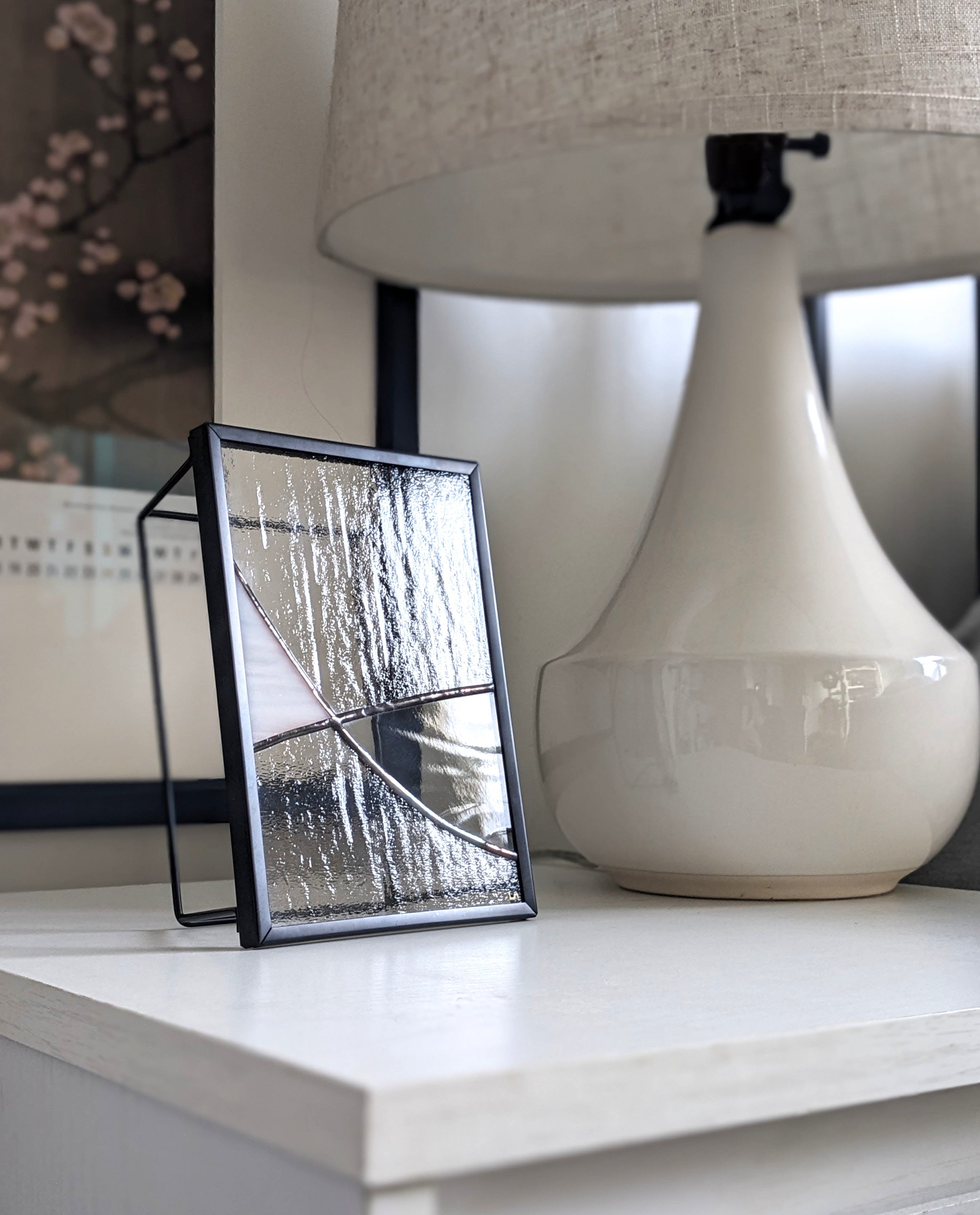 Framed abstract glass art on a shelf with a lamp in the background