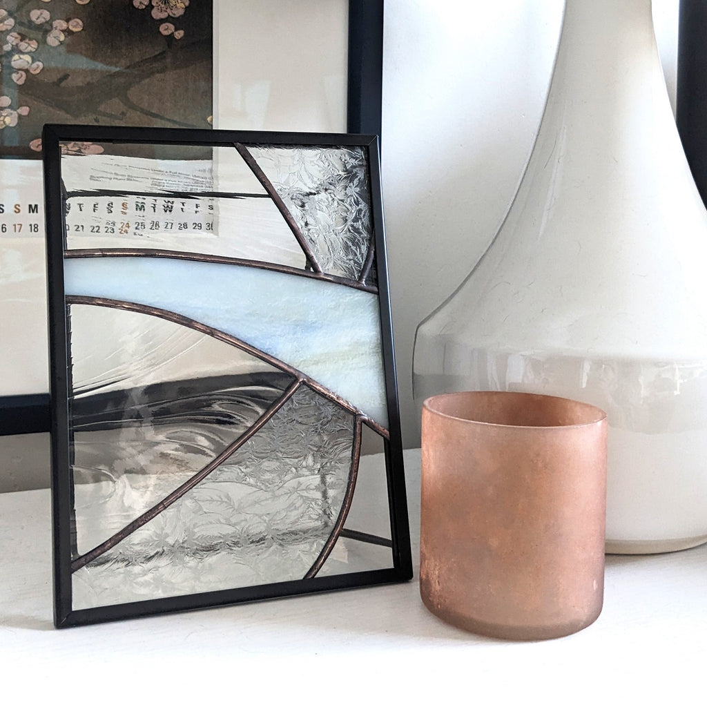 Framed glass artwork with geometric design on a white surface next to a copper cup and a white lamp.