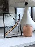 Textured glass artwork with golden yellow detail in modern black frame on a surface with a lamp and candle.